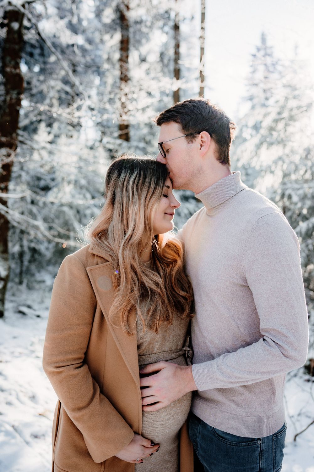 A couple embraces in a snowy winter forest setting wearing warm layers with snow-covered evergreen trees in background.