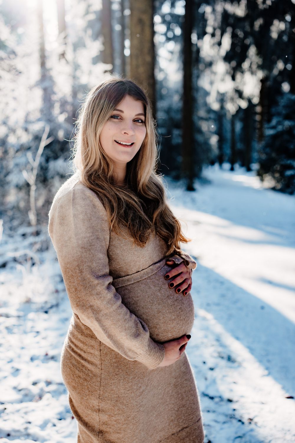 A person wearing a beige sweater poses in a snowy winter forest setting with bright sunlight filtering through trees.