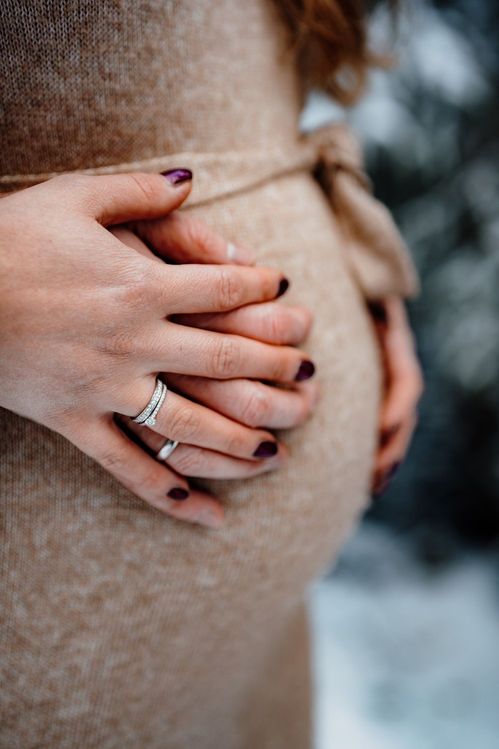Close-up view of hands wearing a diamond engagement ring, with dark burgundy nail polish against a blurred background.