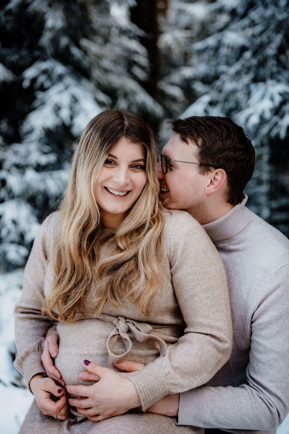 A romantic winter engagement photoshoot with a couple wearing beige sweaters surrounded by snow-covered trees.