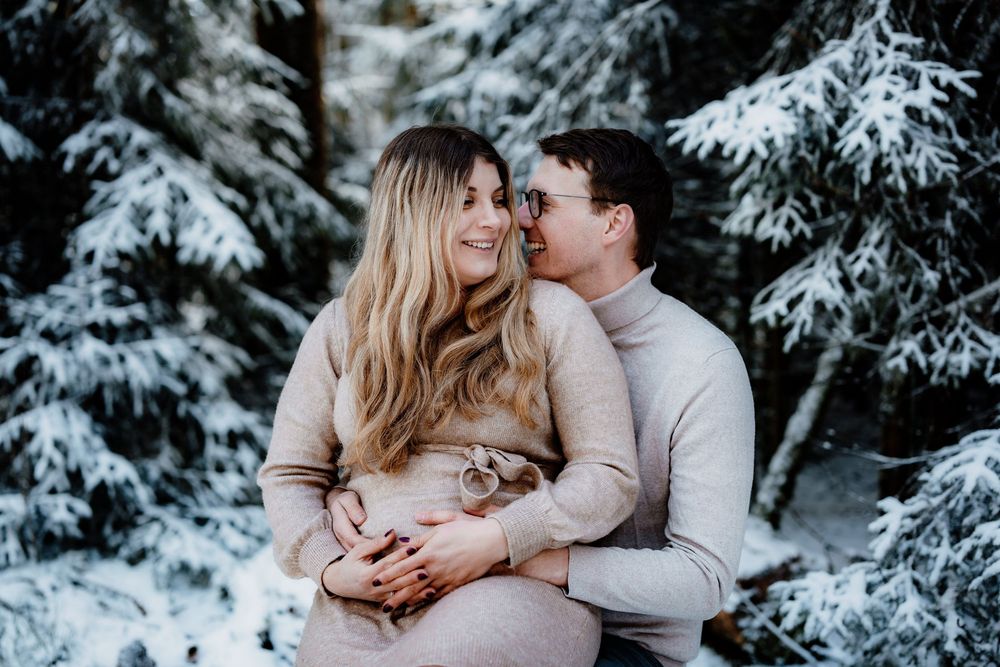 A romantic couple embraces in a snowy winter setting with snow-covered evergreen trees in the background.
