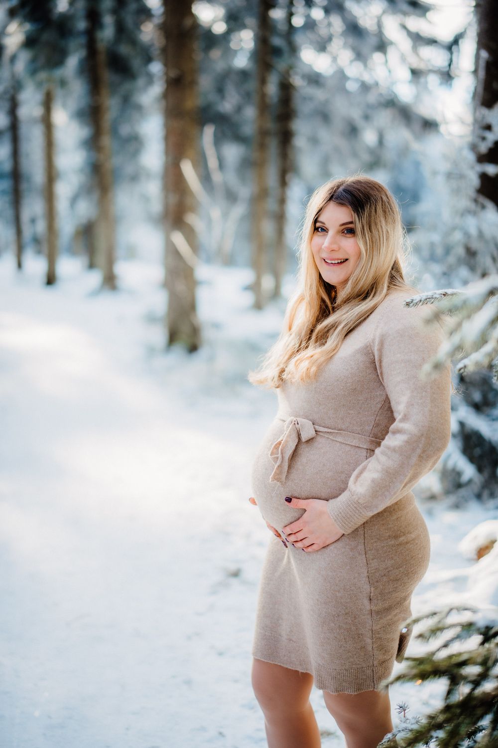 Woman in beige sweater dress stands in snowy winter forest landscape with tall pine trees in background.