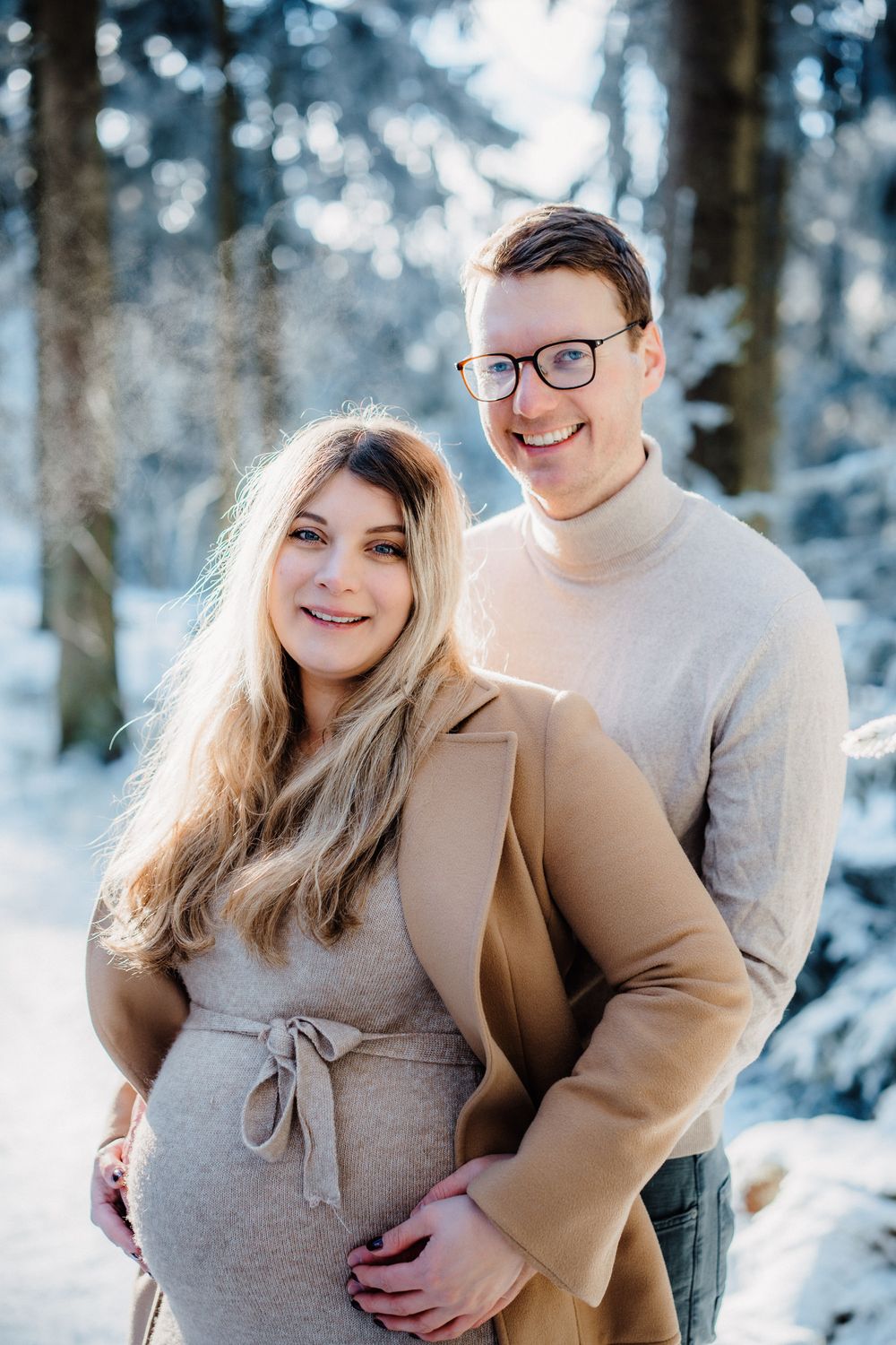 A couple in casual winter attire embraces and smiles in a snowy forest setting.