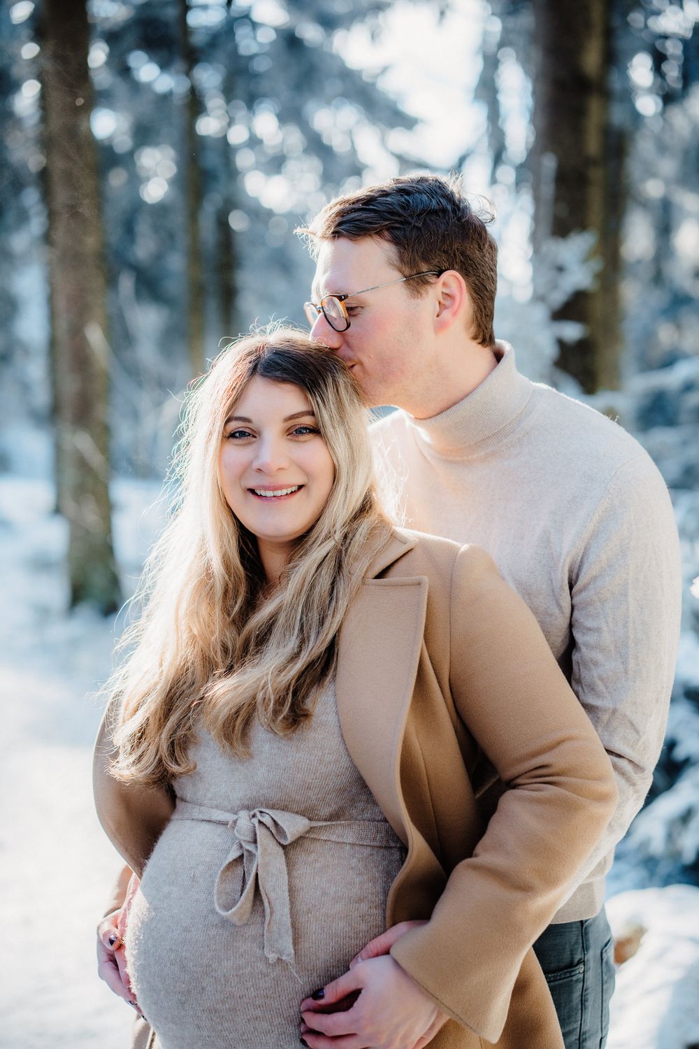 A couple shares an intimate embrace in a snowy winter forest setting while wearing warm neutral-toned clothing.