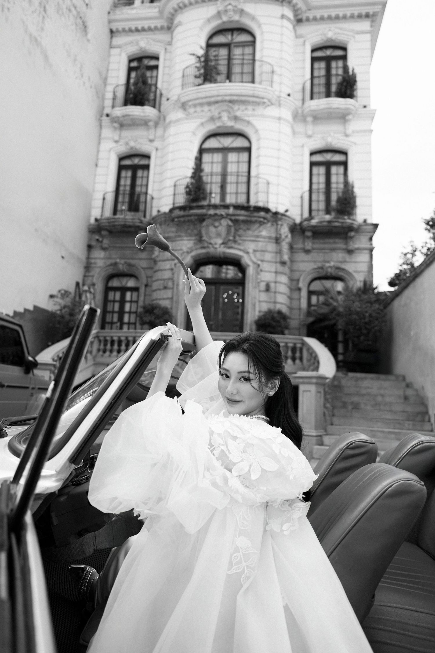 Black and white photo of joyful bride in white dress posing in vintage convertible outside grand building.