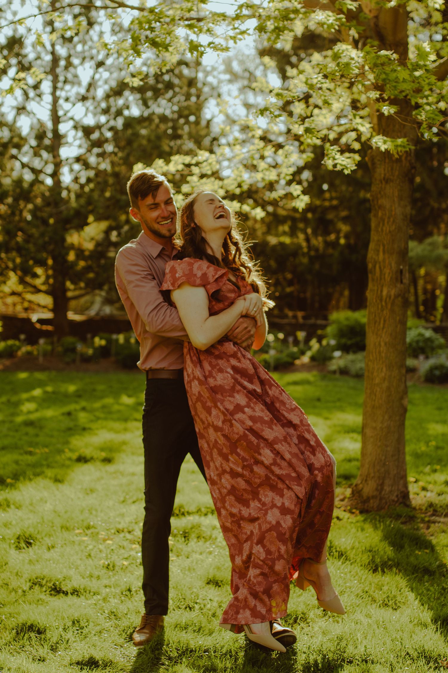 A playful couple dances together on grass beneath trees during golden hour.