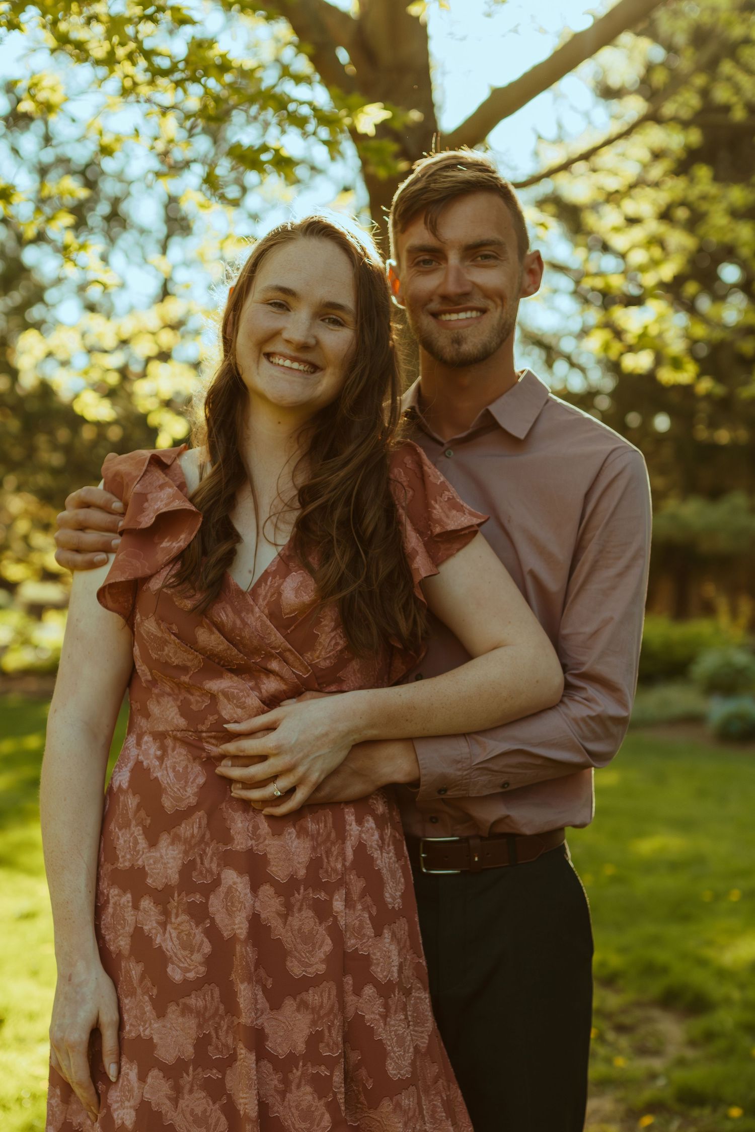 A romantic couple embracing and laughing together in a sun-dappled garden setting.