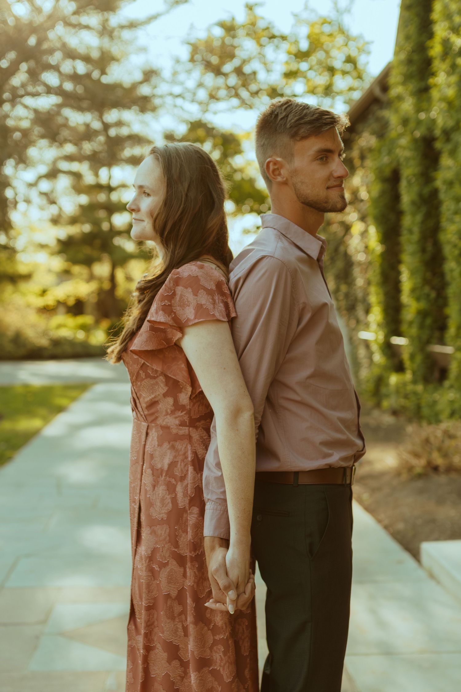 A couple in formal wear stands back-to-back on a paved pathway surrounded by greenery.