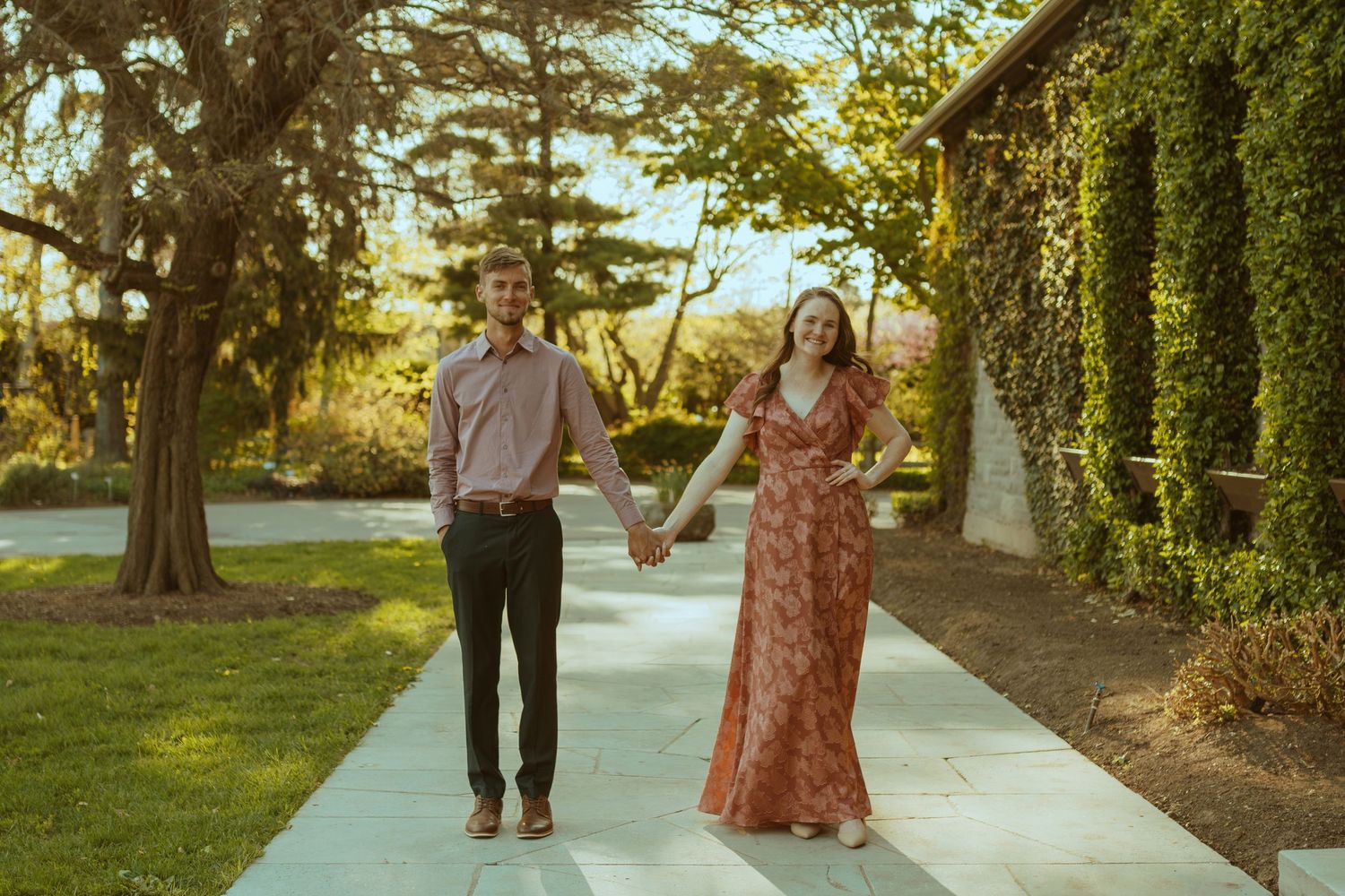 A couple walks together on a peaceful path surrounded by lush green trees and foliage in golden sunlight.