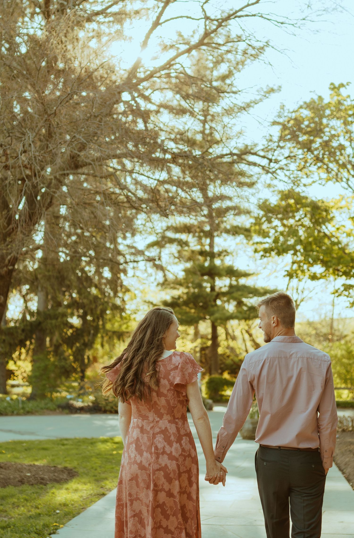 Couple walking hand in hand on a sunlit path with evergreen trees in the background.