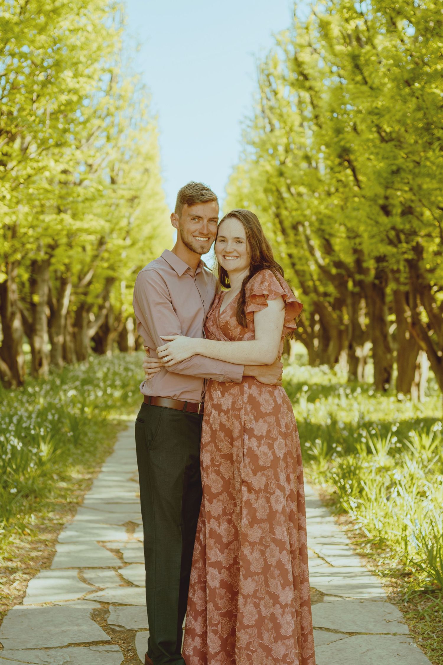 A couple poses together on a tree-lined path during a warm, sunny afternoon.