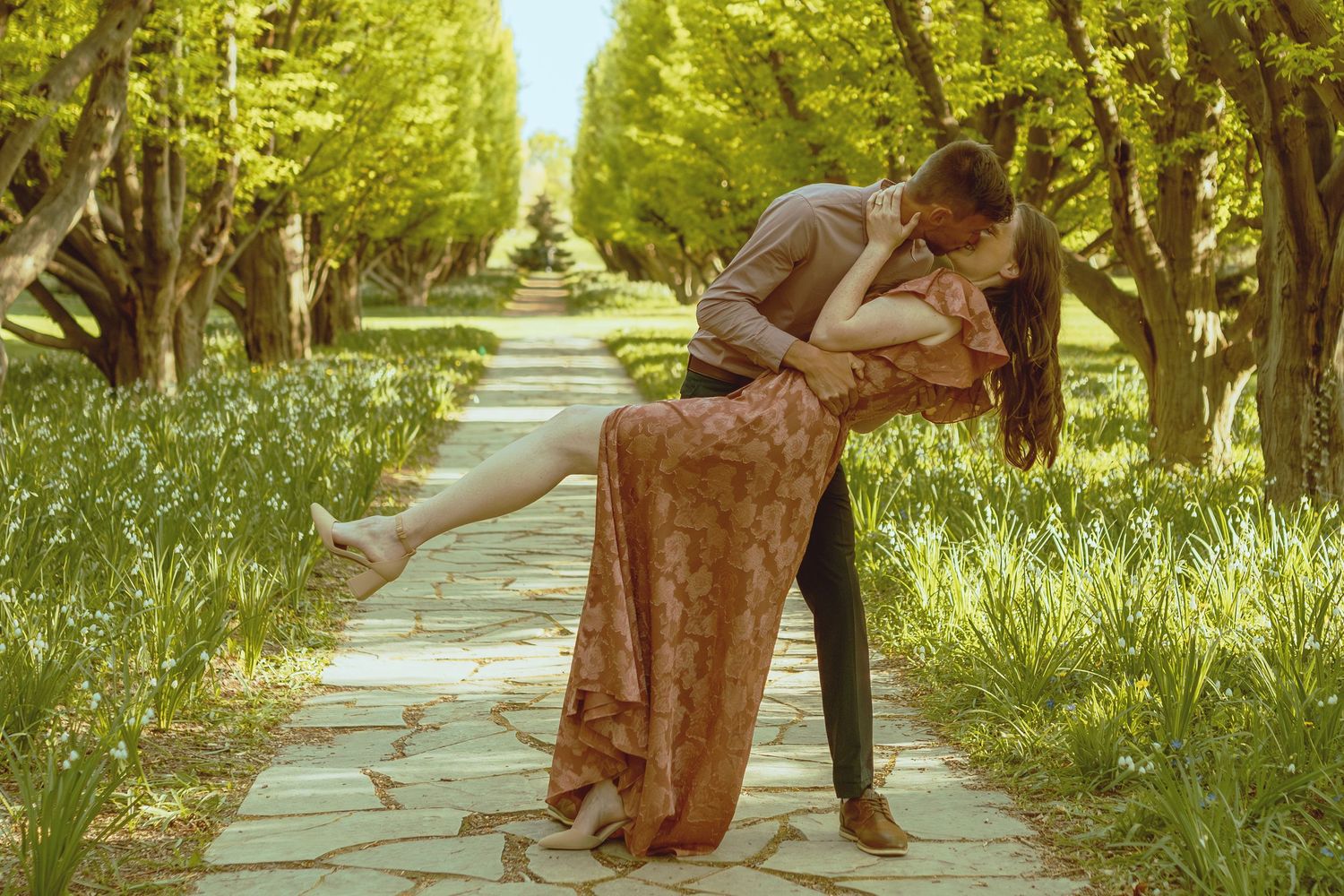 Two people dance together on a sunlit pathway lined with trees and grass.