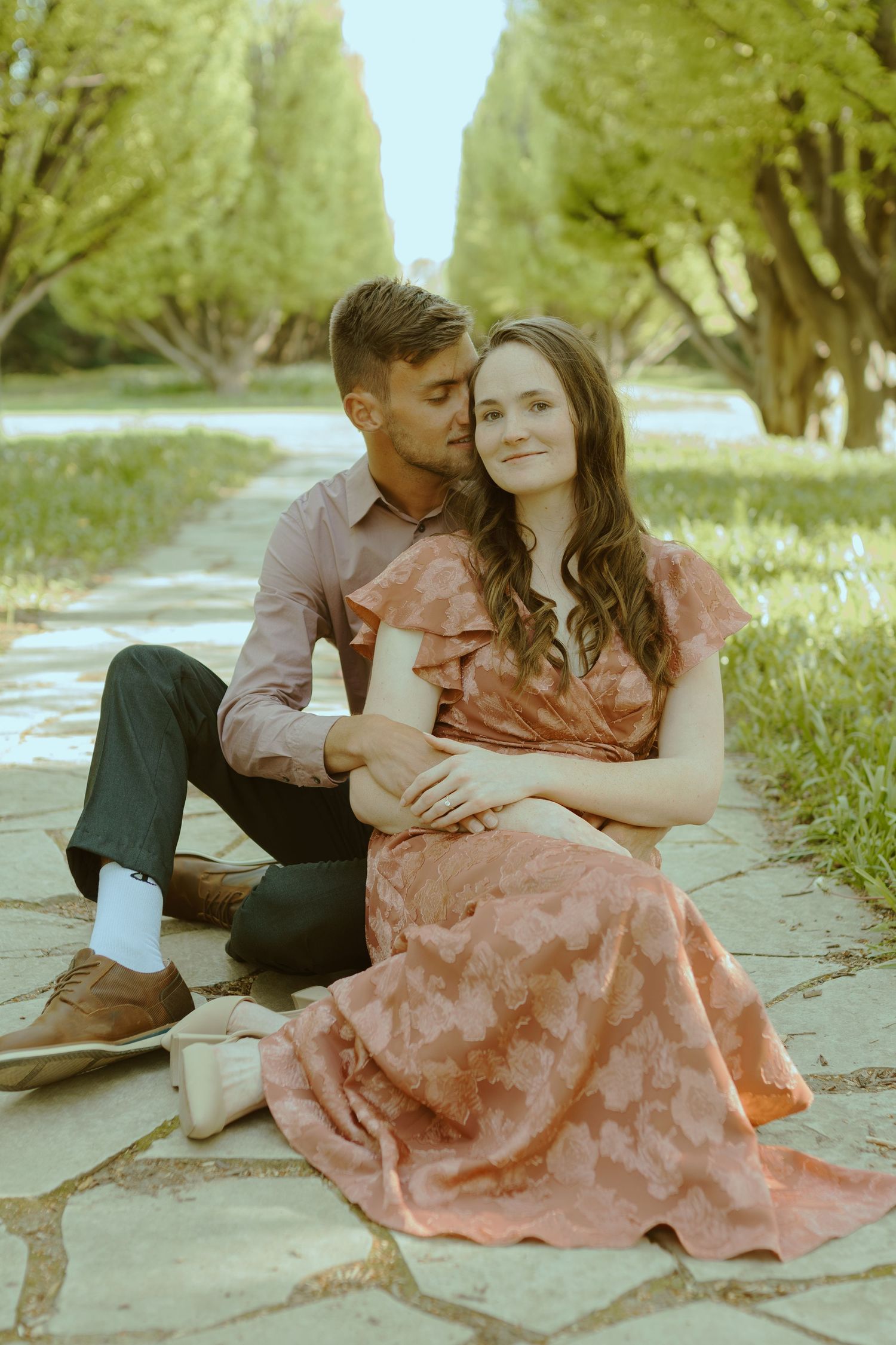 Couple sitting intimately together on stone pavement surrounded by lush greenery on a sunny day.