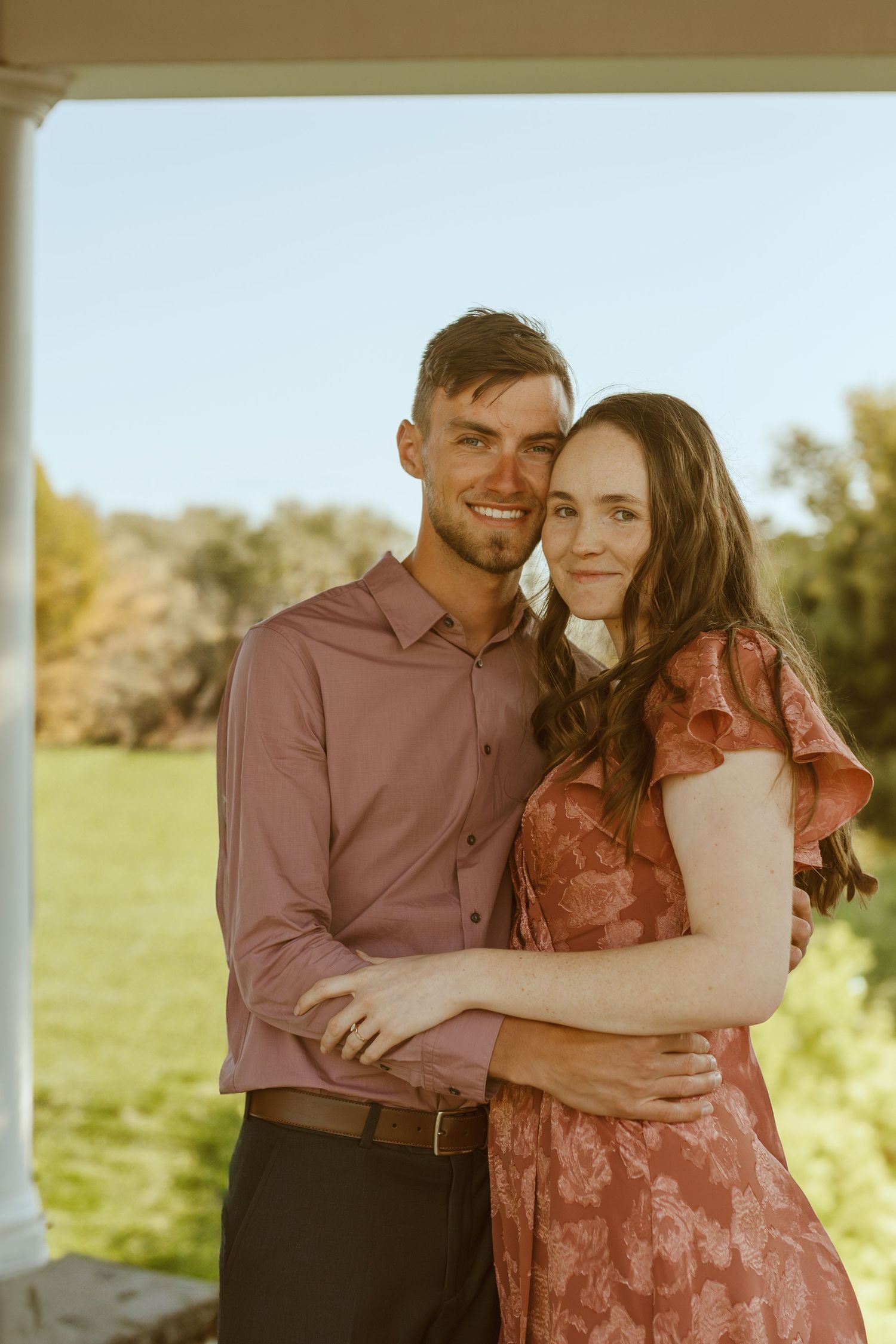 Romantic couple embraces in a sunlit field during golden hour photoshoot in burgundy and pink outfits.