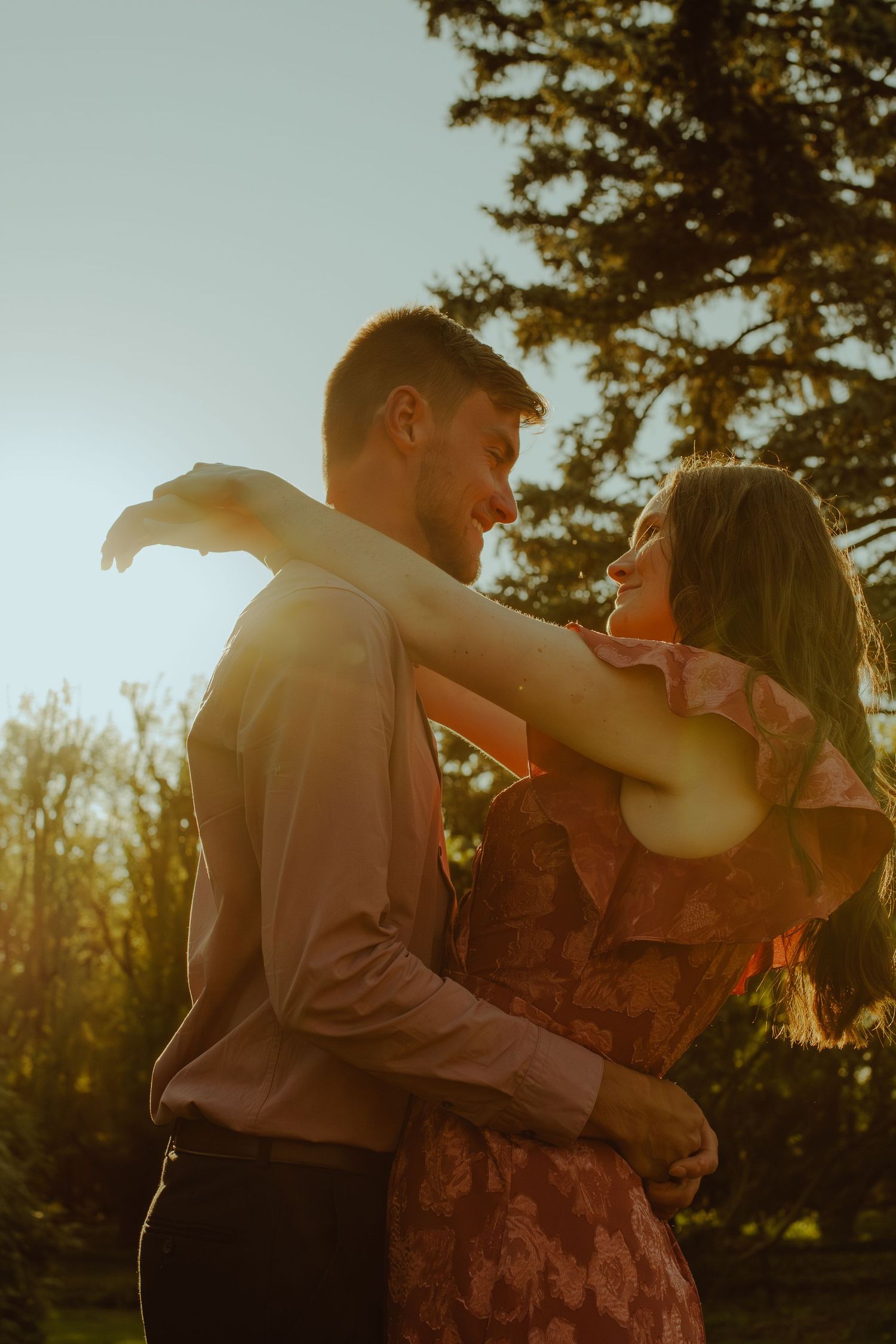 A romantic couple dancing in a golden sunset with trees in the background and warm backlighting creating a silhouette.