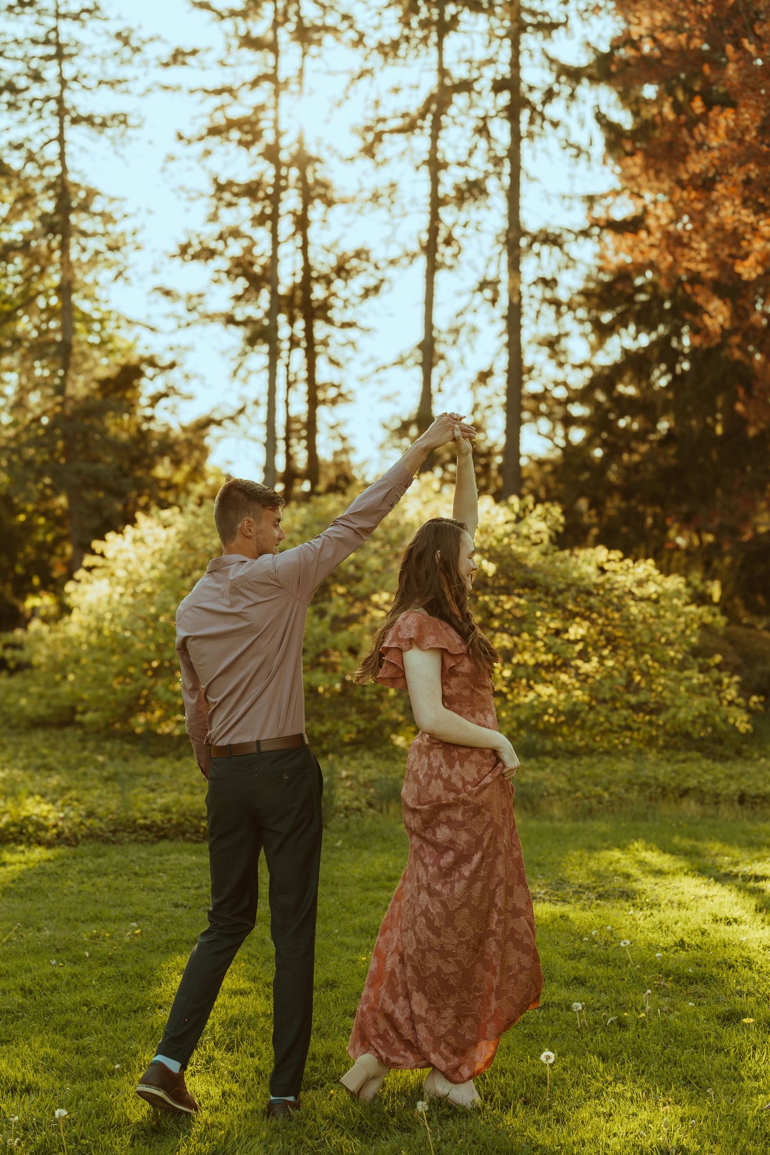 Dancing couple in a grassy meadow with evergreen trees in the background during golden hour.