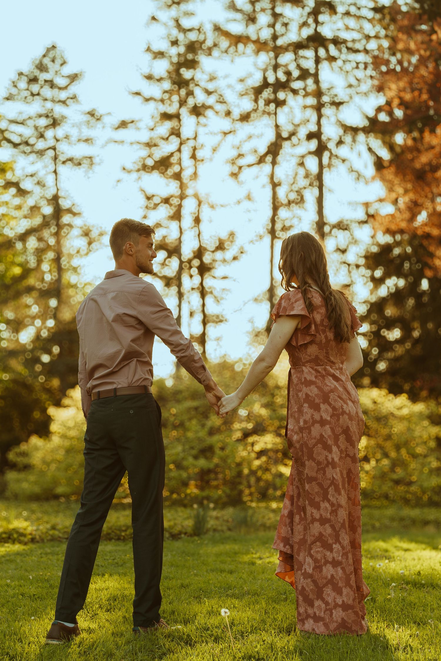 A couple dances playfully on a grassy hill with evergreen trees and warm sunlight creating a magical scene.