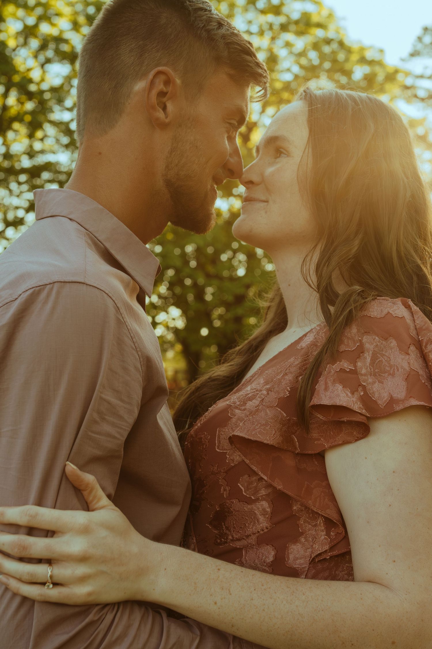 A romantic couple embraces in golden sunlight among trees while wearing formal attire.