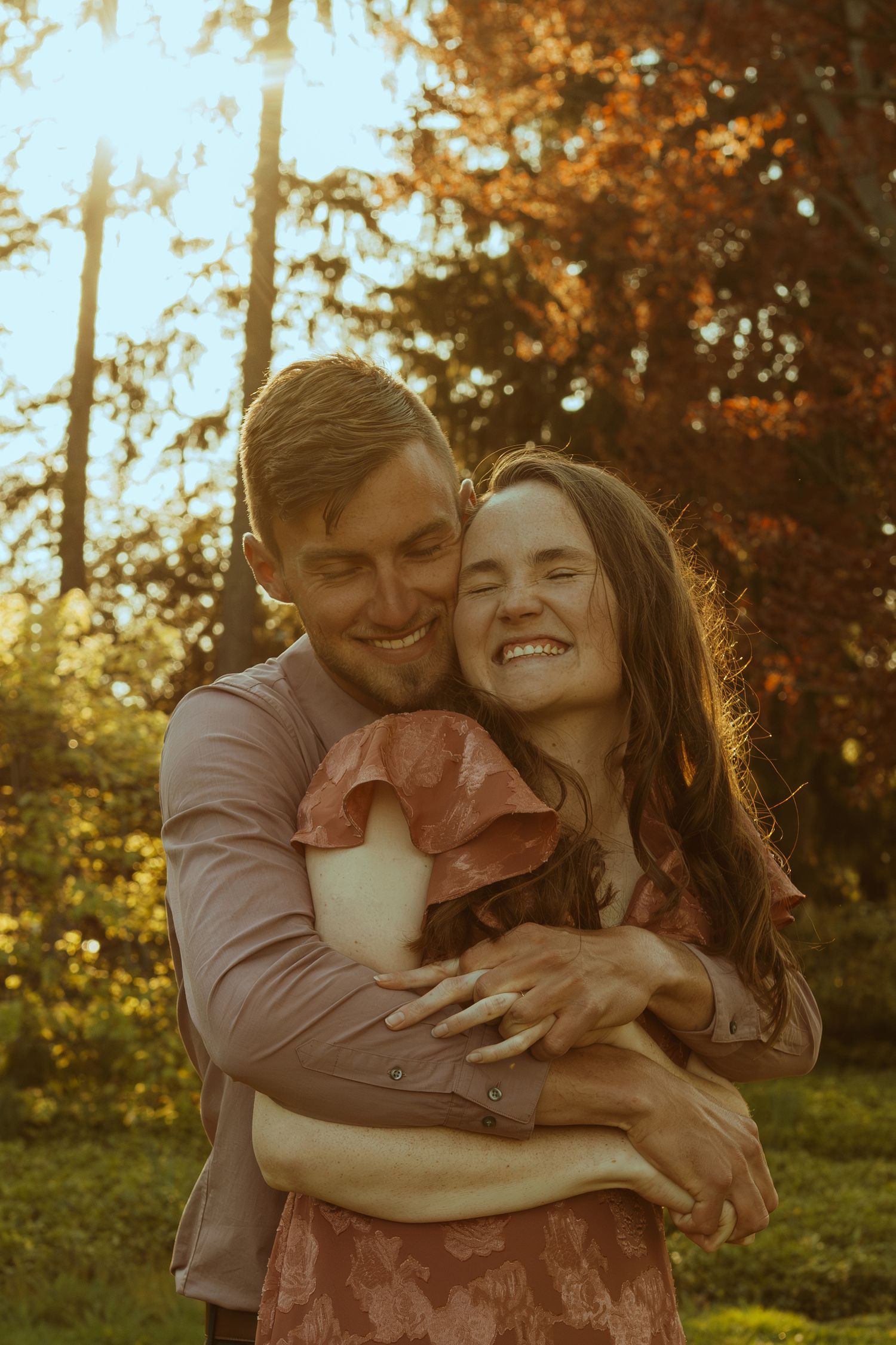 A romantic couple embraces and laughs together in a sunlit outdoor setting with trees in the background.