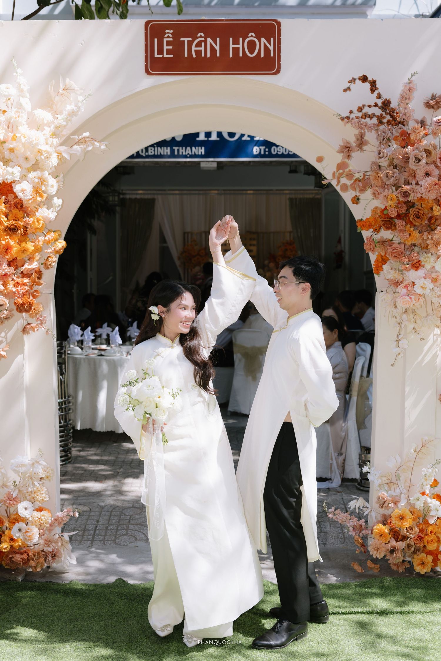 Vietnamese couple in white ao dai dancing under Le Tan Hon arch with autumn floral wedding decor.