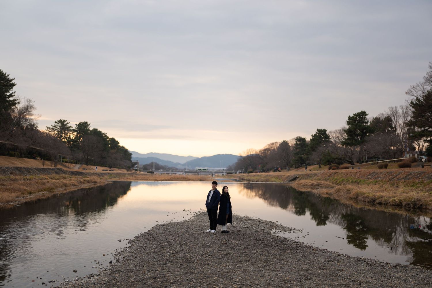 Couple standing on a sandy riverbank with calm water reflections and mountain views at dusk.