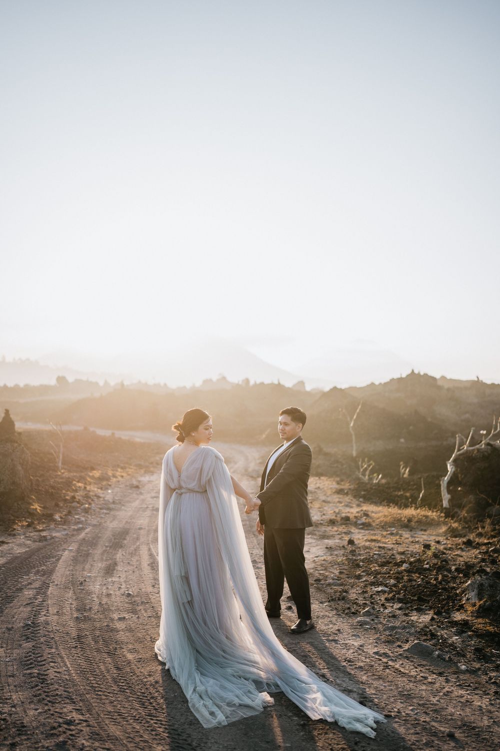 A couple in formal wear walks along a dusty mountain path at sunset.