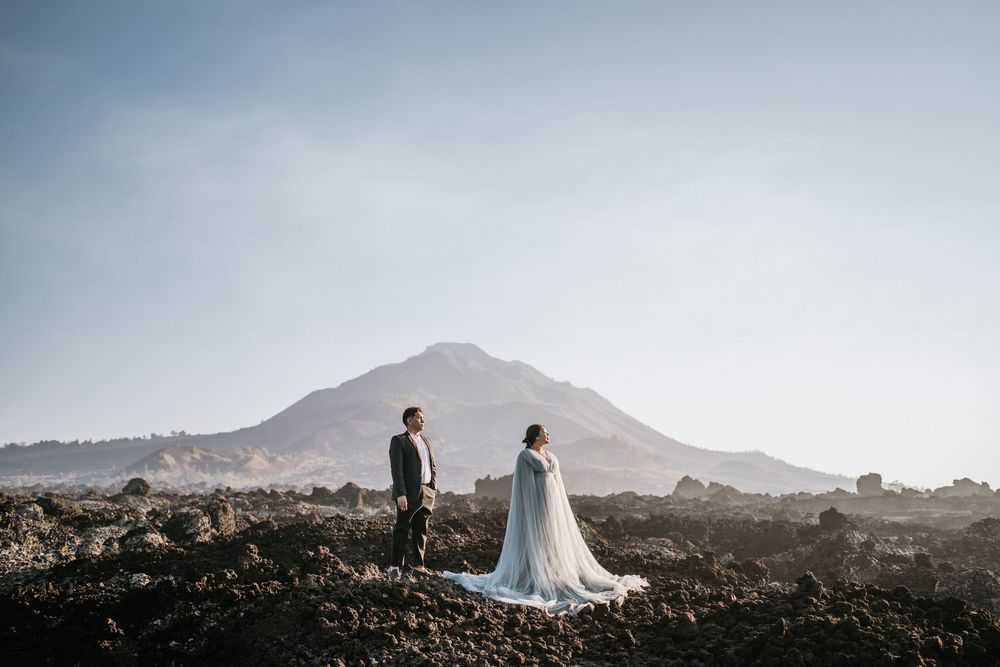 Two people in formal wear stand together on rocky terrain with mountains in background.