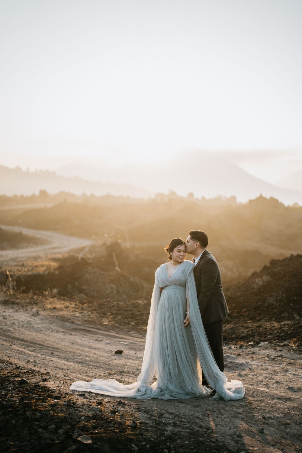 A couple in formal wear sits together on rocky terrain at sunset with mountains in the background.
