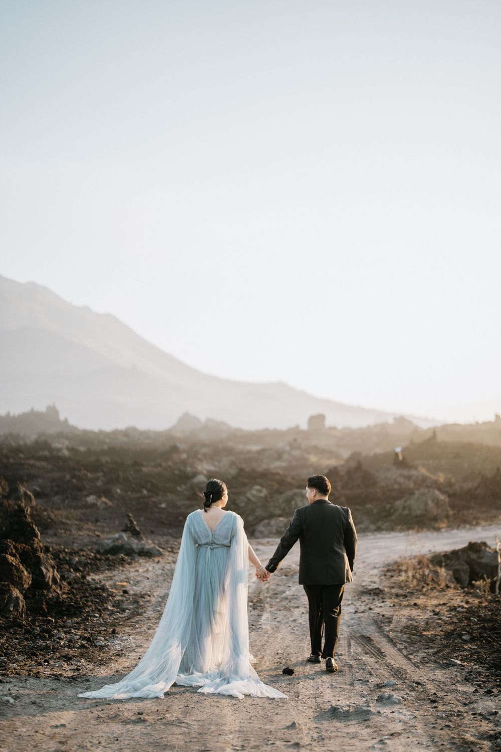 A couple walks hand in hand on a rocky path with mountains and sunset in the distance.