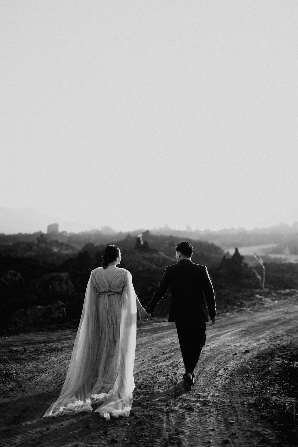 A couple in formal attire walks along a dirt path during golden hour.