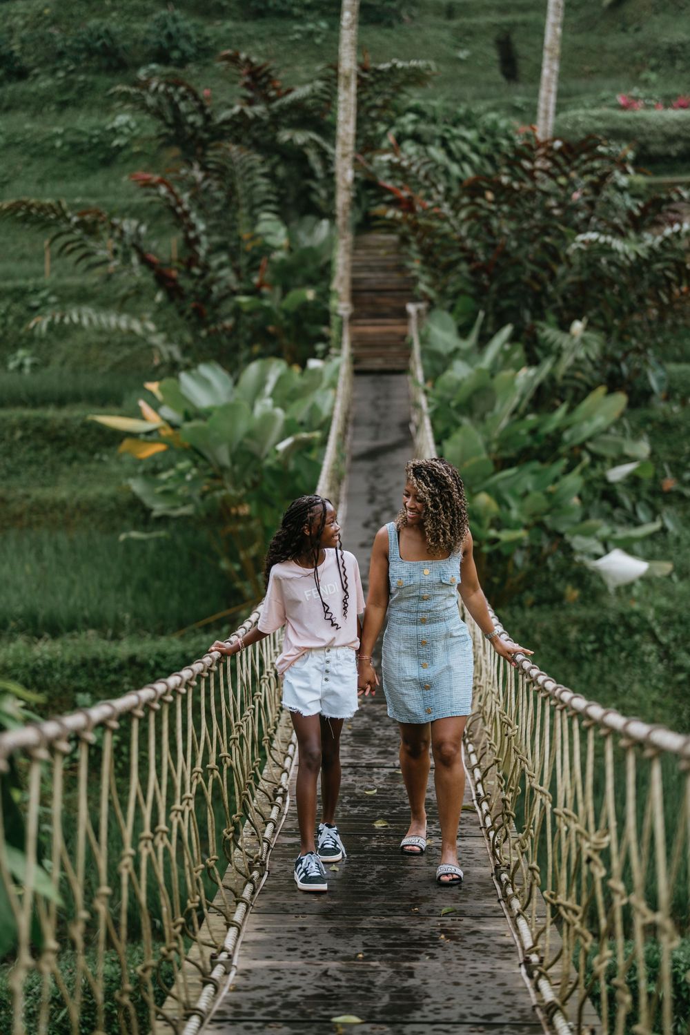 Two people walking across a rope bridge surrounded by lush tropical vegetation.