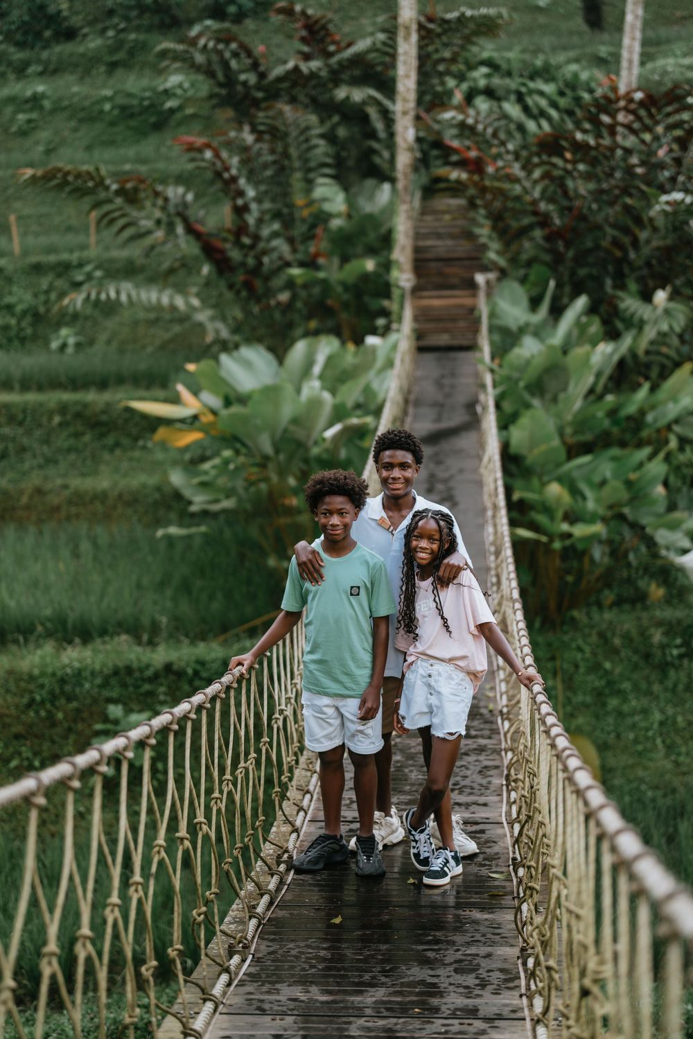 Three people pose together on a rustic rope bridge in a tropical forest setting.
