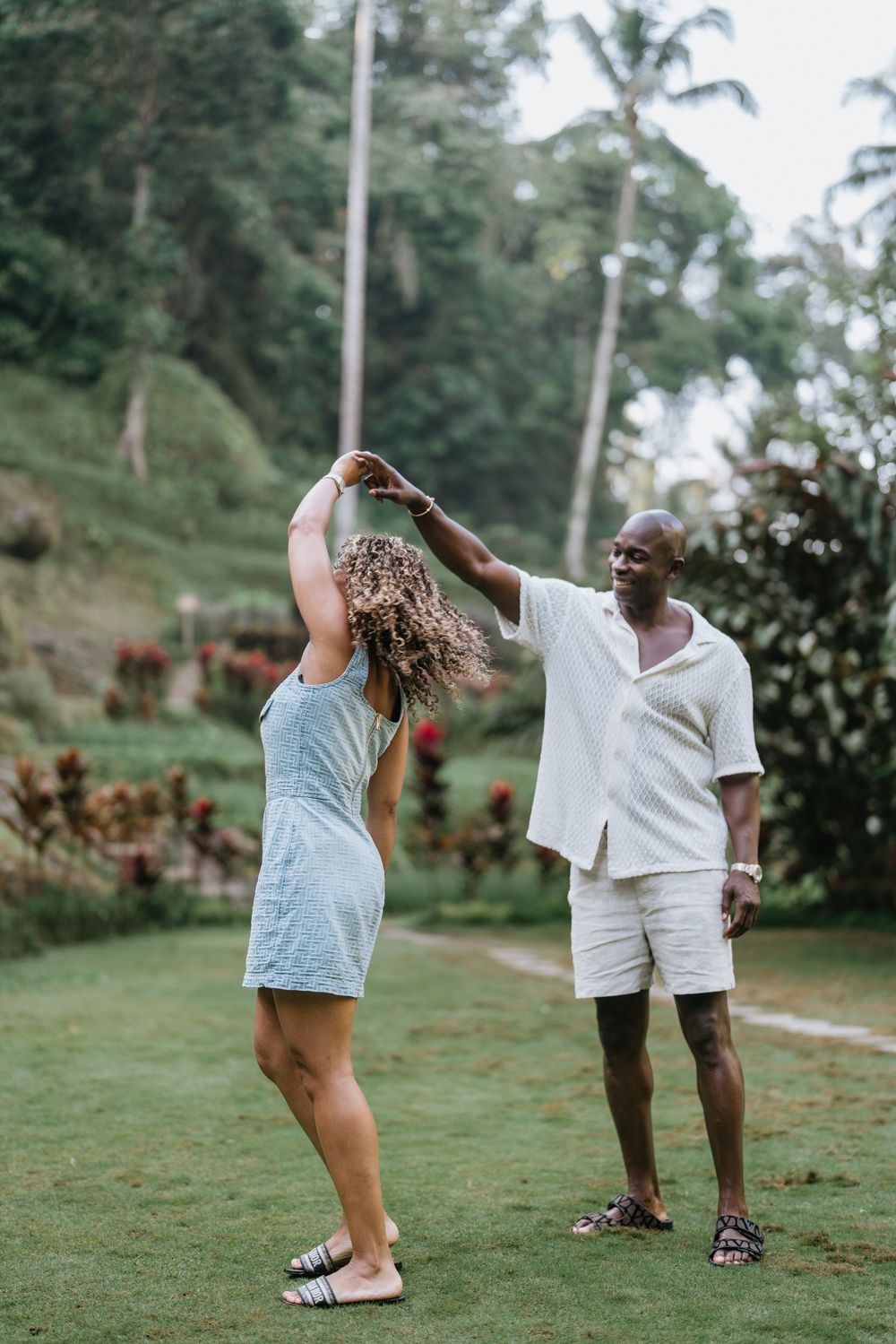 A couple dances playfully on grass surrounded by palm trees and tropical foliage.