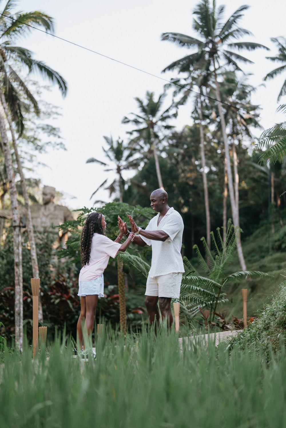 Two people share a playful moment in a grassy field with palm trees in the background.