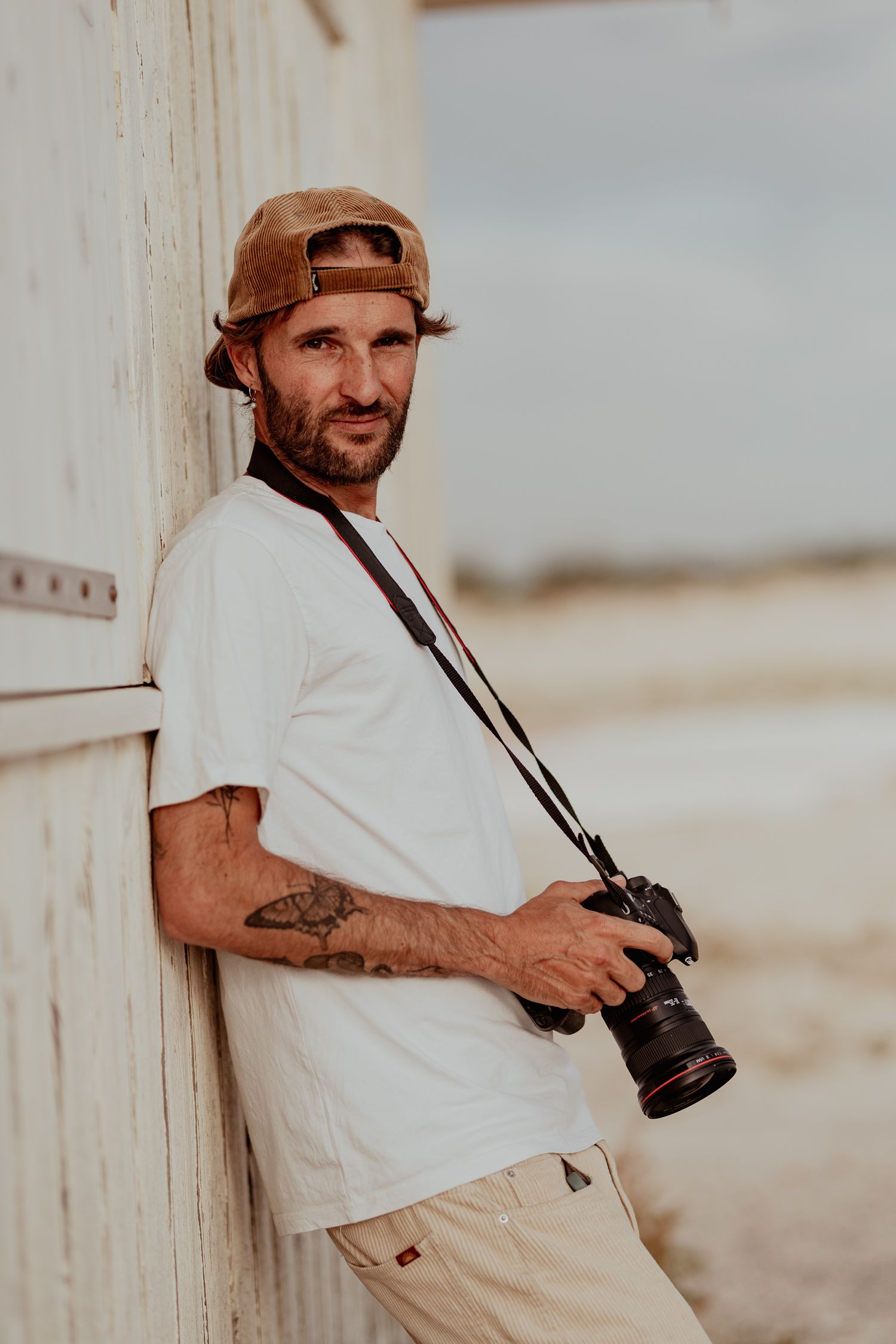 Photographer in white shirt and baseball cap holding camera against beach wall.