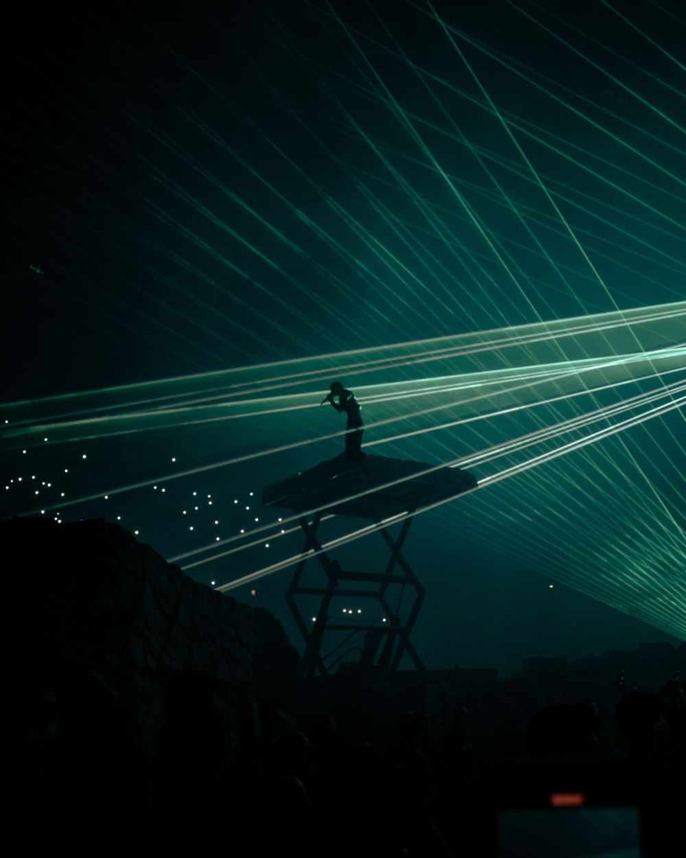 Green laser beams sweep across a dark nighttime sky creating geometric light patterns at a concert venue.