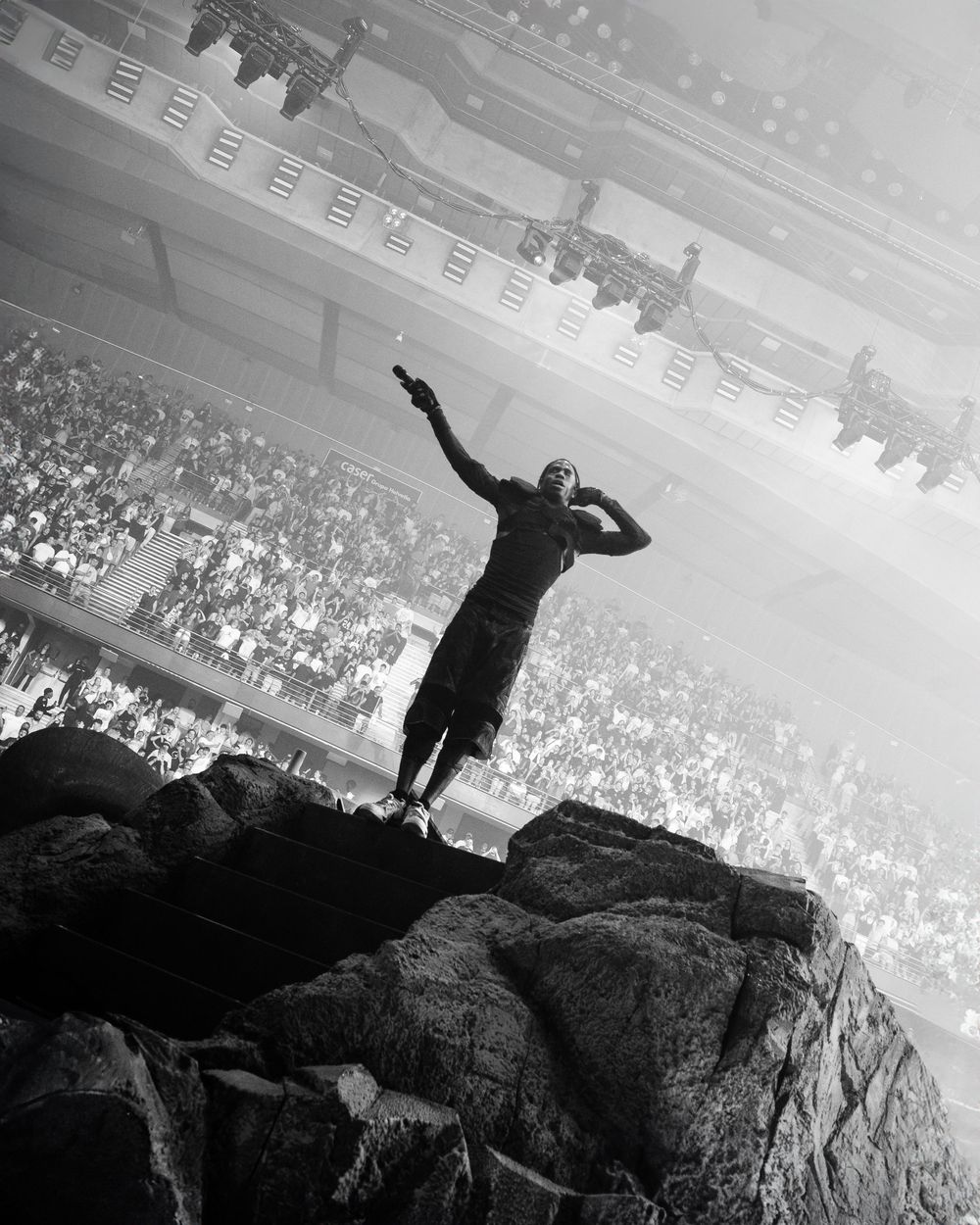 A silhouetted performer stands triumphantly on a rock formation before a large crowd.