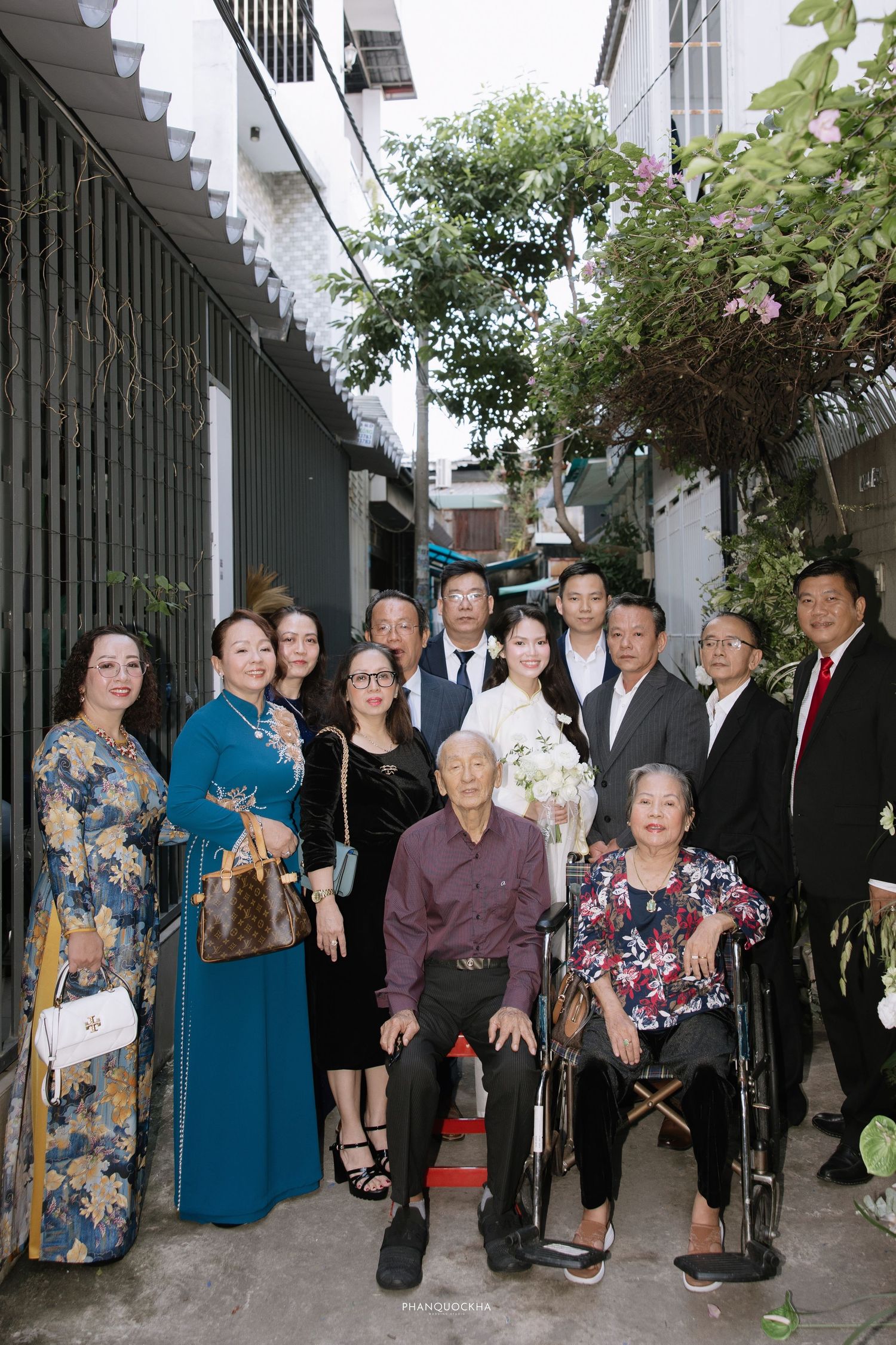 Wedding group photo with bride, groom, and family including elderly seated grandparents outdoors.