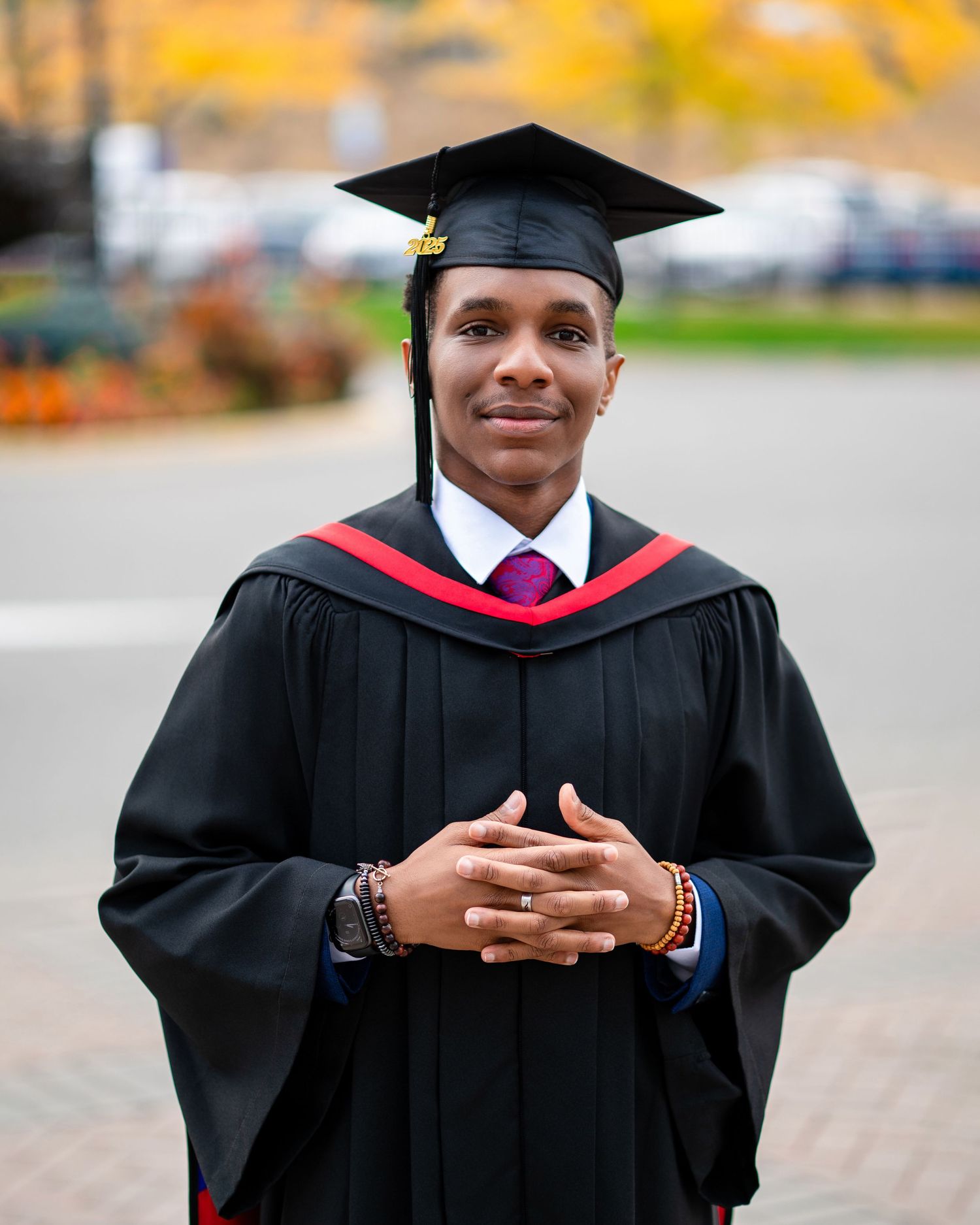 Graduate in black academic regalia poses against lush green foliage.