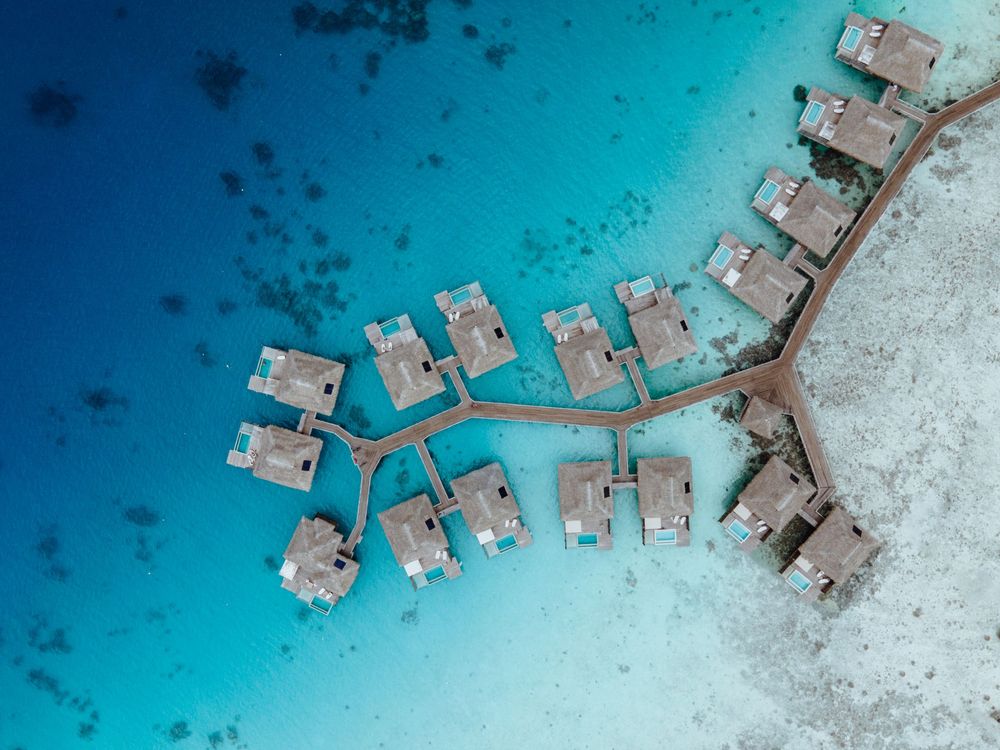 Aerial view of overwater bungalows connected by wooden walkways over crystal clear turquoise lagoon waters.