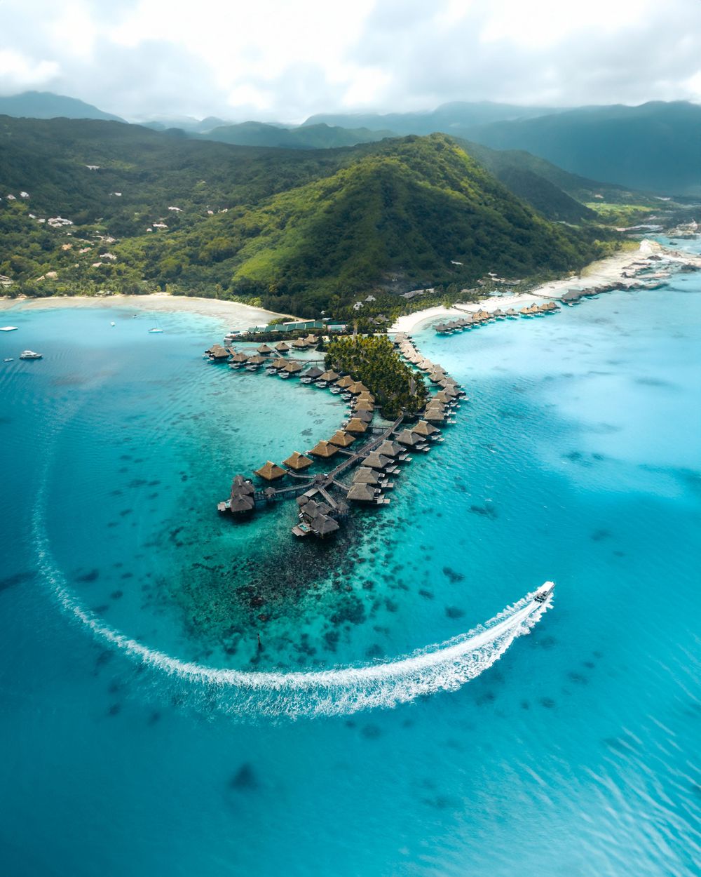 Aerial view of overwater bungalows on a curved peninsula with turquoise waters and a speedboat creating wake in French Polynesia.