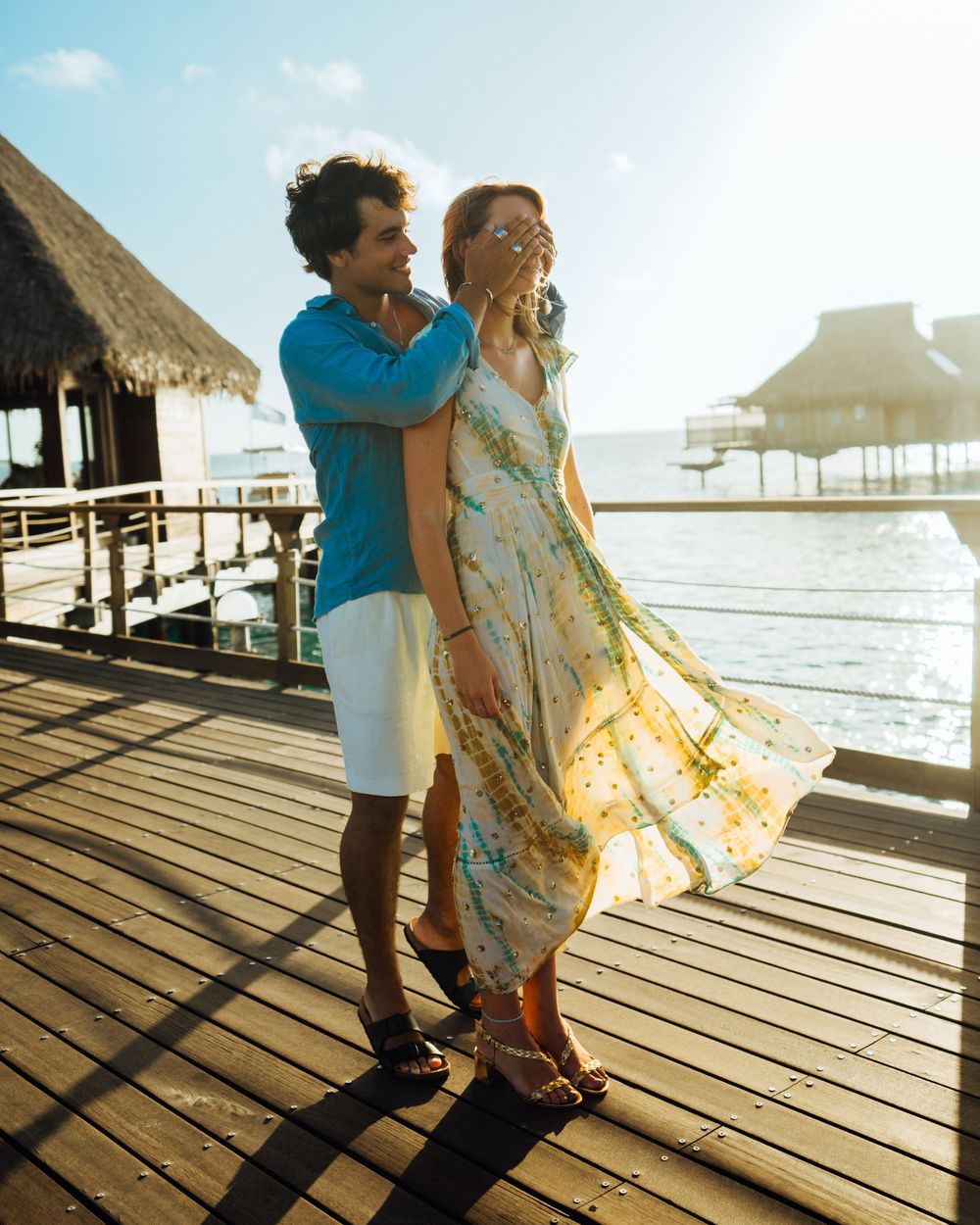 Couple enjoying a romantic moment on a wooden pier at sunset with thatched roof huts and ocean in the background.