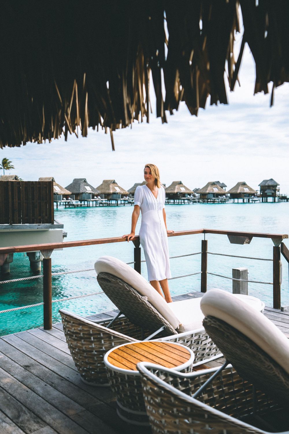 A white-dressed figure stands on an overwater bungalow deck with lounge chairs overlooking turquoise lagoon in the Maldives.