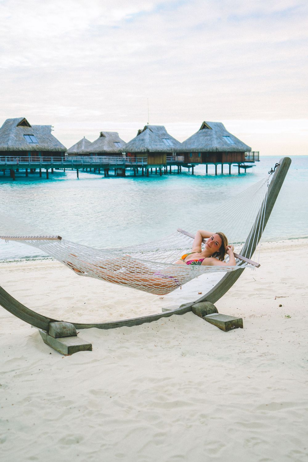 Hammock on a wooden stand overlooking overwater bungalows and turquoise lagoon at a tropical beach resort.