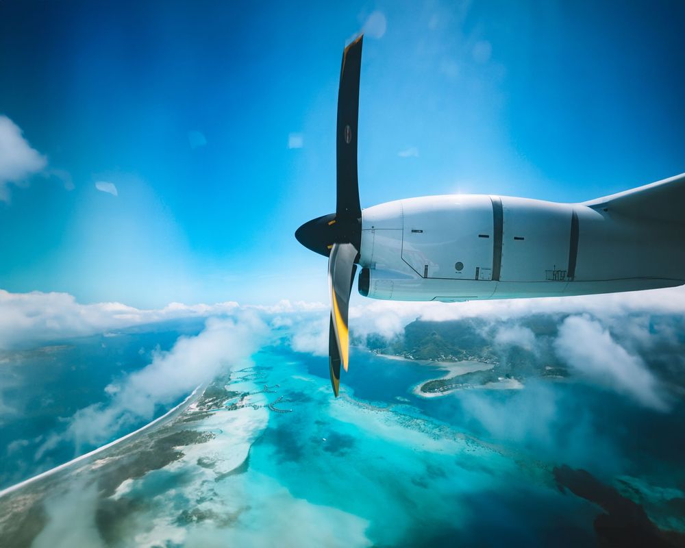 Aerial view from propeller plane window showing turquoise waters and tropical islands below against blue sky.