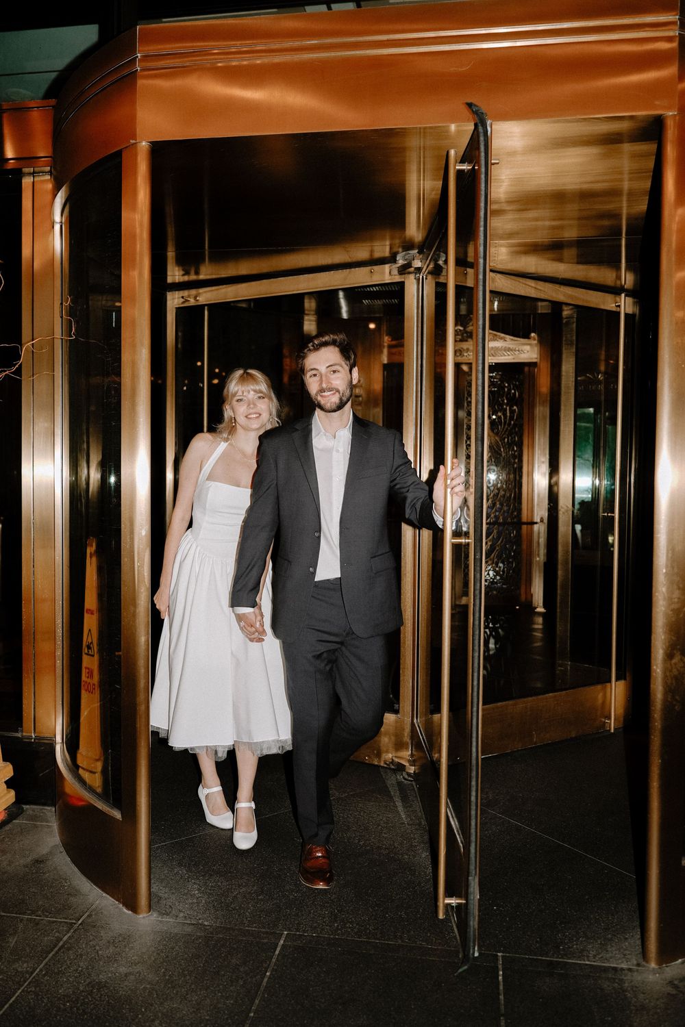 A couple enters a revolving door at night in elegant formal attire.