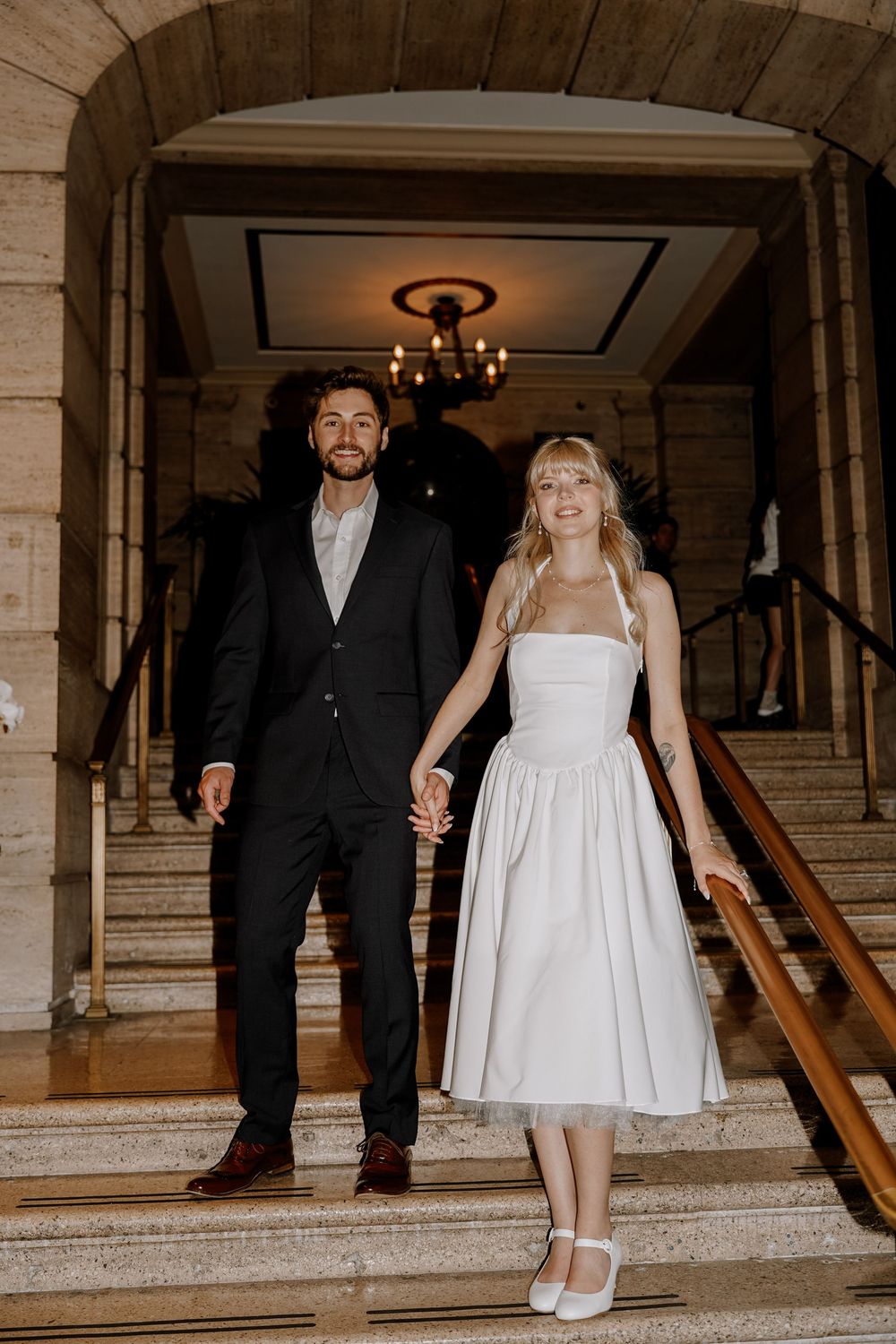 A couple walks down an elegant stone staircase at a formal venue with chandeliers and marble details.