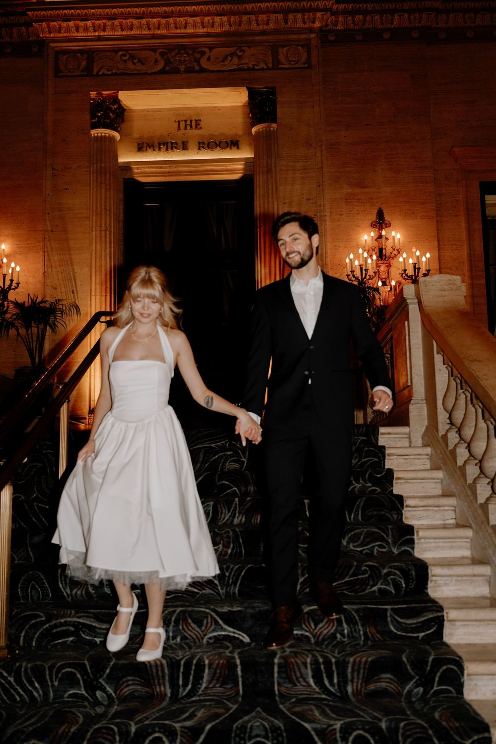 A couple descends a grand marble staircase with surrounding chandeliers and ornate architectural details.