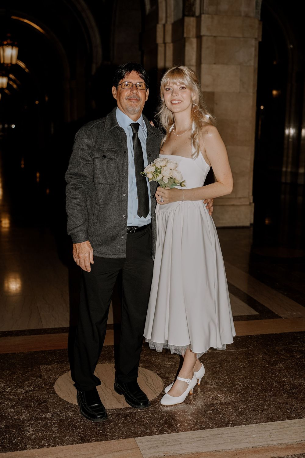 A couple stands in an elegant archway at night, with the bride wearing a white tea-length dress holding a bouquet.