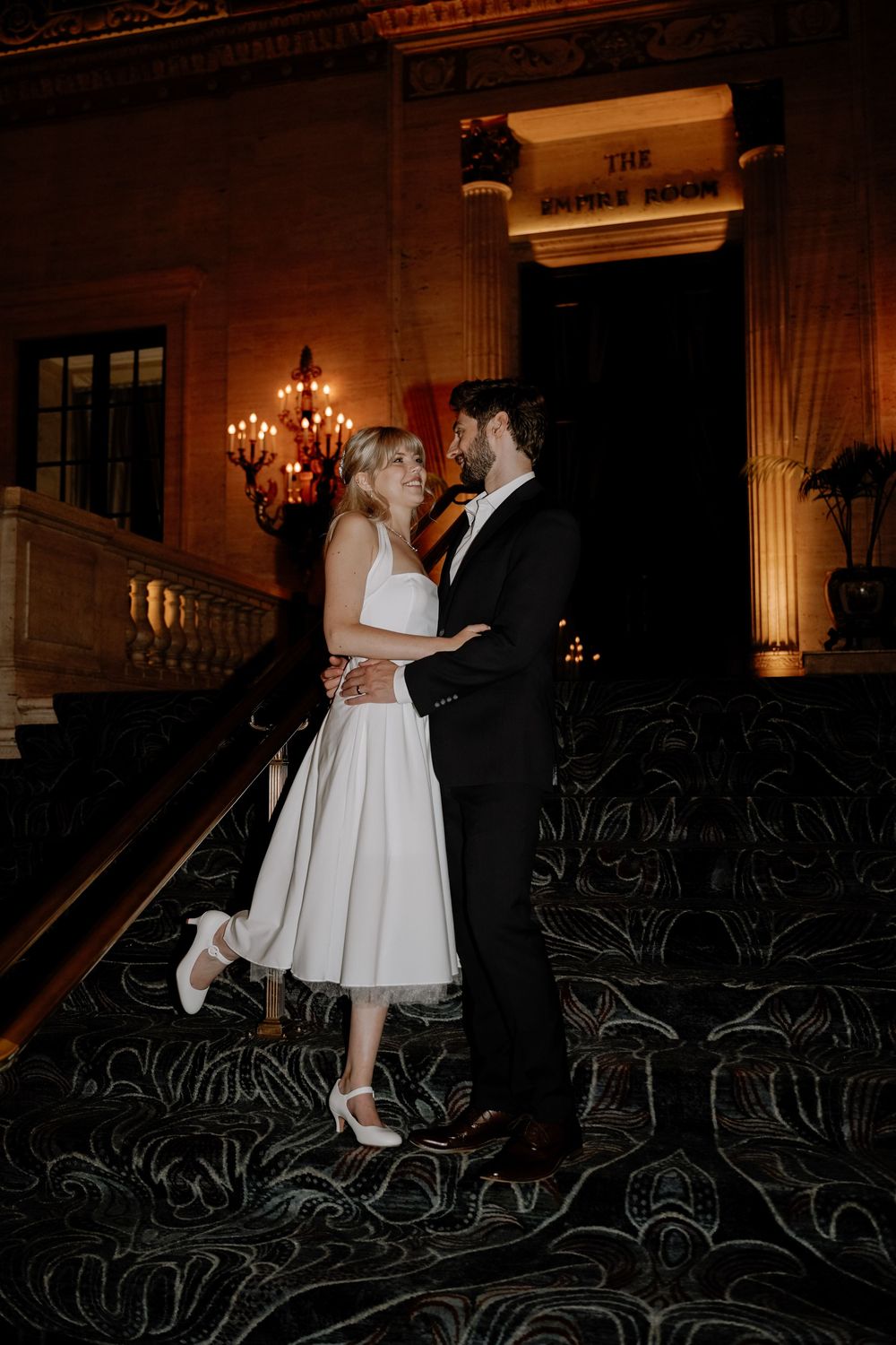 A romantic couple dancing together at a dimly lit venue with elegant chandeliers and dark seating in the background.